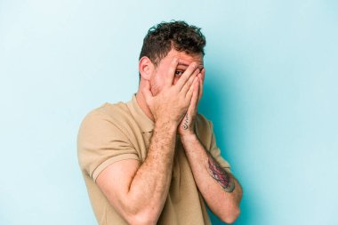 Kafkasyalı genç adam mavi arka planda izole edilmiş. Korkmuş ve gergin parmaklarıyla göz kırp..