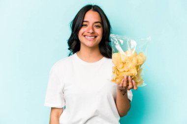 Mavi arka planda izole edilmiş bir paket cipsi tutan genç İspanyol kadın mutlu, güler yüzlü ve neşeli.
