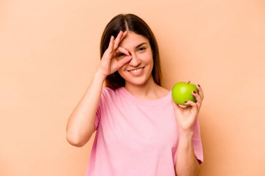 Elinde bej arka planda izole edilmiş bir elma tutan genç İspanyol kadın heyecanlı..