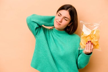 Genç İspanyol kadın elinde bir çanta dolusu cipsle bej arka planda izole edilmiş kafasının arkasına dokunuyor, düşünüyor ve bir seçim yapıyor..
