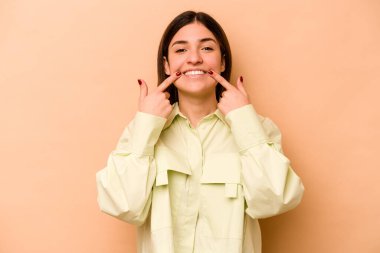 Latin kökenli genç bir kadın bej arka planda izole edilmiş gülümsüyor, parmaklarını ağzına doğrultuyor..