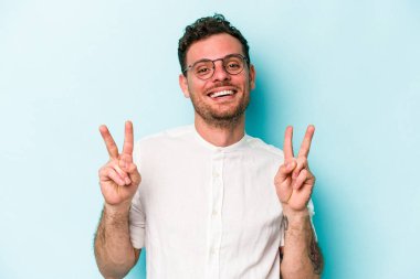 Mavi arka planda izole edilmiş genç beyaz adam zafer işareti gösteriyor ve gülümsüyor..