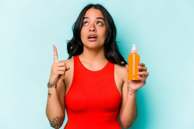 Mavi arka planda güneş kremi tutan genç İspanyol kadın ağzı açık bir şekilde üst tarafı işaret ediyor..