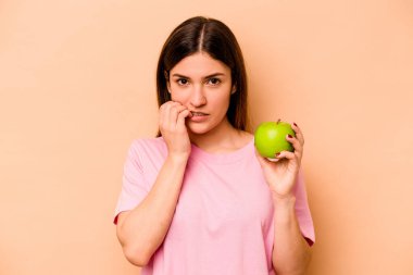 Elma tutan genç İspanyol kadın bej arka planda izole edilmiş tırnak yiyor, gergin ve çok endişeli..