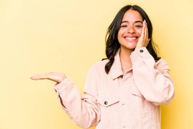 Sarı arka planda izole edilmiş genç İspanyol kadın avuç içinde fotokopi alanı tutuyor, el üstünde tutuyor. Şaşırmış ve mutlu..