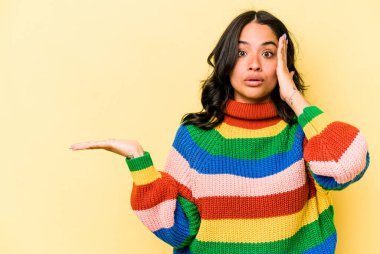 Sarı arka planda izole edilmiş genç İspanyol kadın avuç içi fotokopi alanını tutmaktan etkilenmiş..