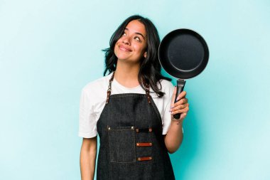 Young hispanic cooker woman holding frying pan isolated on blue background dreaming of achieving goals and purposes