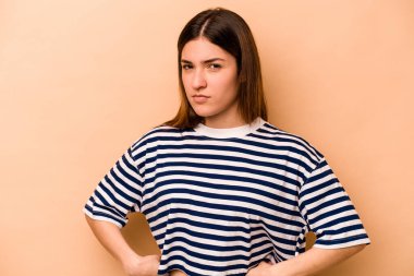 Latin kökenli genç bir kadın bej arka planda izole edilmiş hoşnutsuzca kaşlarını çatmış, kollarını kavuşturmuş..