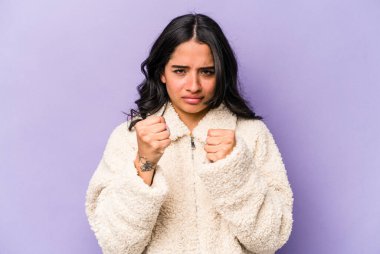 Mor arka planda izole edilmiş genç İspanyol kadın kameraya yumruğunu, agresif yüz ifadesini gösteriyor..