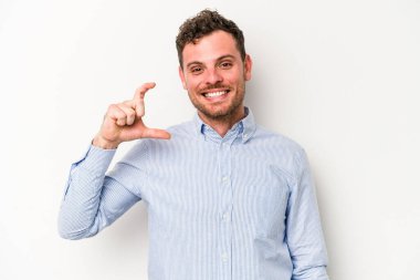 Beyaz arka planda izole edilmiş genç beyaz bir adam işaret parmaklarıyla küçük bir şey tutuyor, gülümsüyor ve kendinden emin..