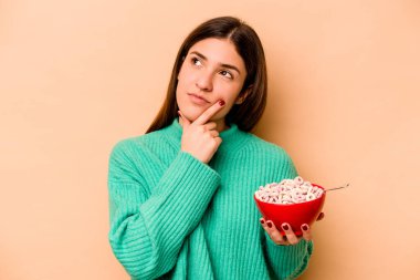 Latin kökenli genç bir kadın bej arka planda izole edilmiş mısır gevreği yiyor. Şüpheli ve şüpheci bir ifadeyle yana bakıyor..