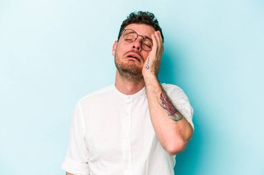 Kafkasyalı genç adam mavi arka planda izole edilmiş. Yorgun ve uykulu. Elini başının üstünde tutuyor..