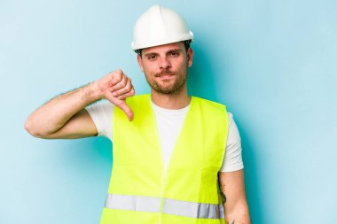 Genç, beyaz bir adam mavi arka planda izole edilmiş. Sevimsiz bir jest gösteriyor. Başparmakları aşağıda. Anlaşmazlık konsepti.