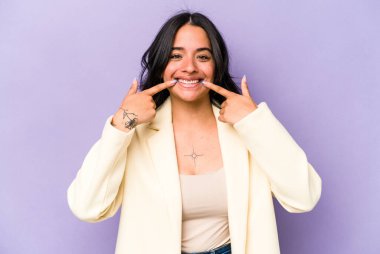 Mor arka planda izole edilmiş genç İspanyol kadın gülümsüyor, parmaklarını ağıza doğrultuyor..