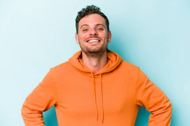 Kafkasyalı genç, mavi arka planda izole edilmiş. Kendine güvenen, kalçaları tutan biri..