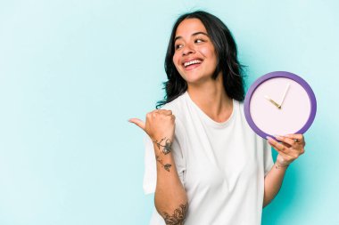 Mavi arka planda bir saat tutan genç İspanyol kadın parmak uzakta, gülerek ve kaygısız.