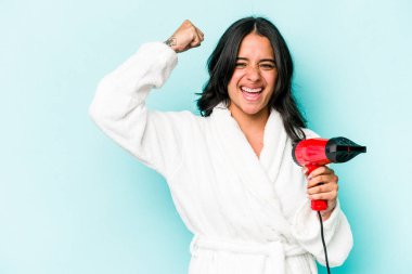 Mavi arka planda izole edilmiş kurutma makinesini tutan genç İspanyol kadın zaferden sonra yumruğunu kaldırıyor, kazanan konsept..