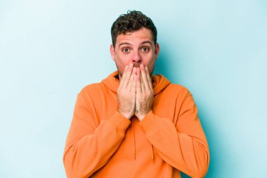 Mavi arka planda izole edilmiş genç beyaz adam şok olmuş, ağzı ellerle kaplanmış, yeni bir şey keşfetmeye hevesli..