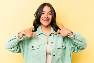 Sarı arka planda izole edilmiş genç İspanyol kadın parmağını göstererek şaşırdı, geniş bir gülümsemeyle..