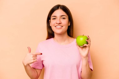 Elma tutan genç İspanyol kadın, bej arka planda izole edilmiş bir şekilde gömlek fotokopi alanını işaret ediyor, gururlu ve kendinden emin.