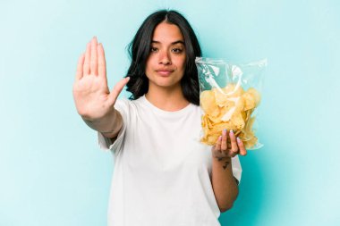 Mavi arka planda izole edilmiş bir paket cipsi tutan genç İspanyol kadın elini uzatmış, dur işaretini göstererek seni engelliyor..