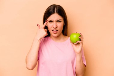 Elinde bej arka planda izole edilmiş bir elma tutan genç İspanyol kadın işaret parmağıyla hayal kırıklığı jesti gösteriyor..