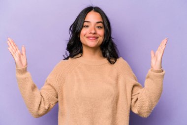 Mor arka planda izole edilmiş genç İspanyol kadın işaret parmaklarıyla küçük bir şey tutuyor, gülümsüyor ve kendinden emin..