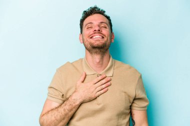 Mavi arka planda izole olmuş genç beyaz adam yüksek sesle gülüyor ve elini göğsüne koyuyor..