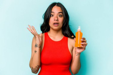 Mavi arka planda güneş kremi tutan genç İspanyol kadın şaşırdı ve şok oldu..