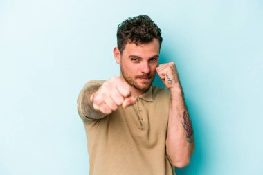Kafkasyalı genç bir adam mavi arka planda izole edilmiş. Yumruk atıyor, sinirleniyor, kavga ediyor, kavga ediyor..