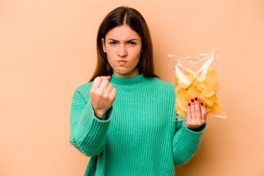 Bej arka planda izole edilmiş bir çanta dolusu cipsi tutan genç İspanyol kadın kameraya yumruğunu gösteriyor, agresif bir yüz ifadesi..