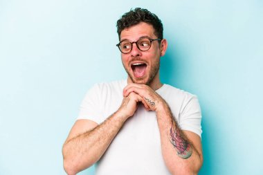 Mavi arka planda izole edilmiş genç beyaz adam ellerini çenesinin altında tutuyor, mutlu bir şekilde yana bakıyor..