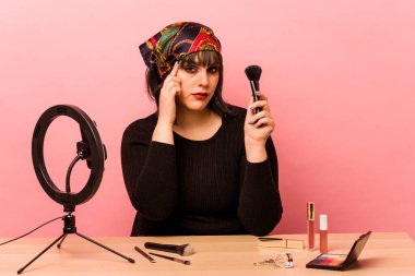 Makyaj eğitmeni genç bir kadın pembe arka planda izole edilmiş bir makyaj dersi veriyor. Şakağı işaret ediyor, düşünüyor, bir göreve odaklanıyor..