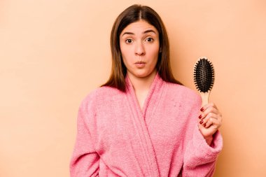 Elinde saç fırçası olan genç İspanyol kadın bej arka planda izole edilmiş omuzlarını silkiyor ve gözleri açık..