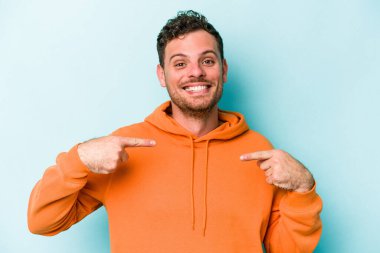 Mavi arka planda izole edilmiş genç beyaz adam parmağını göstererek şaşırdı, geniş bir gülümsemeyle..