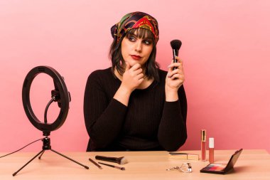 Makyaj sanatçısı genç bir kadın pembe arka planda izole edilmiş şüpheli ve şüpheci bir ifadeyle makyaj dersi veriyor..