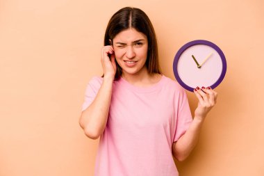 Elinde bej arka planda izole edilmiş bir saat tutan genç İspanyol kadın kulaklarını elleriyle kapatıyor..