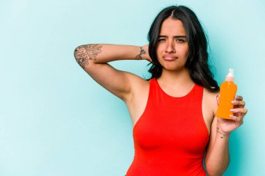 Mavi arka planda güneş kremi tutan genç İspanyol kadın kafasının arkasına dokunuyor, düşünüyor ve bir seçim yapıyor..