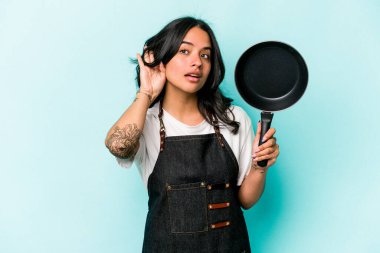 Young hispanic cooker woman holding frying pan isolated on blue background trying to listening a gossip.