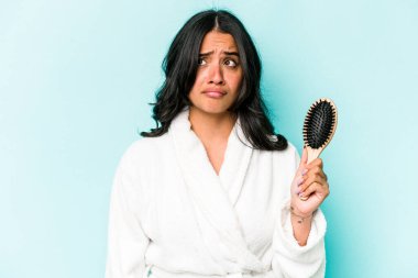 Mavi arka planda saç fırçası tutan genç İspanyol kadın şaşkın ve kararsız hissediyor..