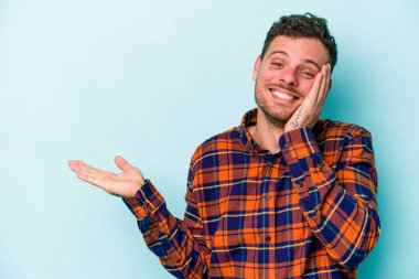 Mavi arka planda izole edilmiş genç beyaz bir adam avucunun içinde fotokopi alanı tutuyor, el üstünde tutuyor. Şaşırmış ve mutlu..