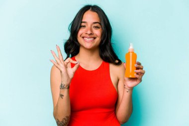 Mavi arka planda güneş kremi tutan genç İspanyol kadın neşeli ve kendinden emin..