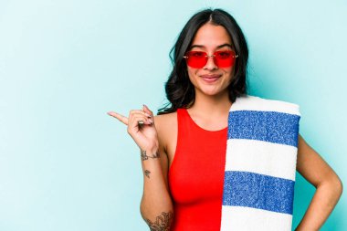 Mavi arka planda izole edilmiş plaj havlusu tutan genç İspanyol kadın gülümsüyor ve boş alanda bir şey gösteriyor..