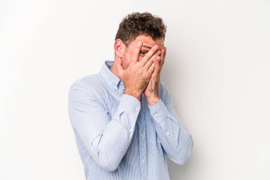 Beyaz arka planda izole edilmiş genç beyaz bir adam parmaklarının arasından korkmuş ve gergin bir şekilde göz kırpıyor..