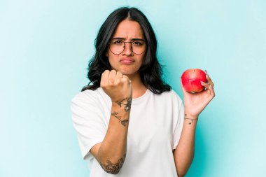 Mavi arka planda izole edilmiş bir elma tutan genç İspanyol kadın kameraya yumruğunu gösteriyor, agresif yüz ifadesi..