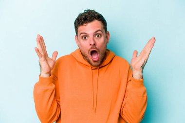 Mavi arka planda izole edilmiş genç beyaz bir adam zaferi ya da başarıyı kutluyor, şaşırıyor ve şok oluyor..