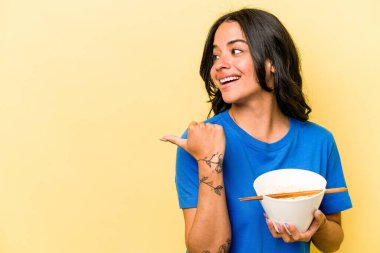 Mavi arka plan noktalarında ramen yiyen genç İspanyol kadın başparmak uzakta, gülüyor ve kaygısız.