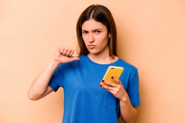 Bej arka planda izole edilmiş cep telefonu tutan genç beyaz kadın gururlu ve kendinden emin hissediyor, örnek olarak.