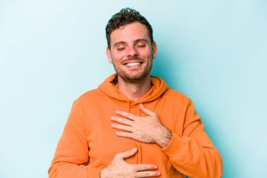 Mavi arka planda izole edilmiş genç beyaz adam mutlu bir şekilde gülüyor ve ellerini karnının üstünde tutarak eğleniyor..