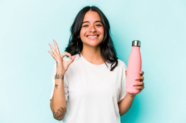 Mavi arka planda termosu olan genç İspanyol kadın neşeli ve kendinden emin..